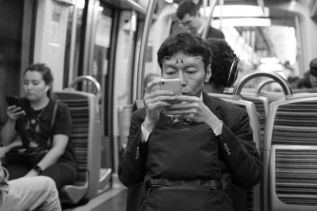 Black and white photograph of a man in a suit looking at his phone while sitting on public transit, with other commuters visible in the background.