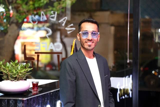 Young man wearing a gray blazer and light blue tinted sunglasses smiling confidently while standing in a cafe. A small potted succulent plant and a glass of red tea sit on the dark counter beside him. Reversed text reading "cafe" is visible on the window behind him.