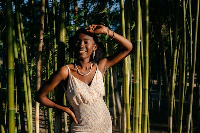 Young Black woman smiling radiantly while standing among tall green bamboo stalks in natural sunlight. She wears a light colored patterned dress and has one hand raised to shield her eyes from the sun, with a pearl necklace visible.
