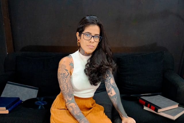 Woman with long dark hair, glasses, and tattoos on her arms wearing a cream top and orange pants, sitting on a dark couch surrounded by books.