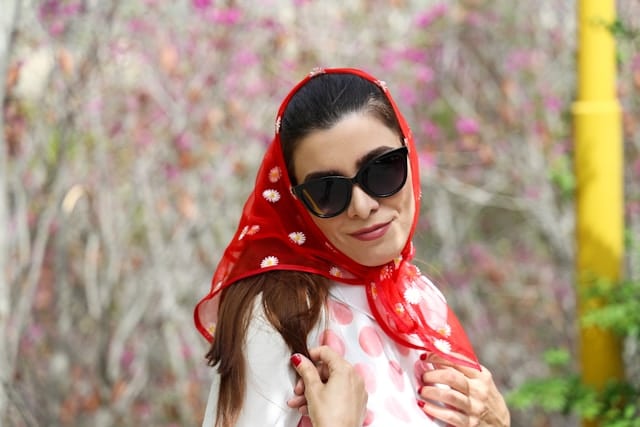 Woman wearing dark sunglasses and a bright red headscarf decorated with small white flowers, with a subtle smile on her face. She has long brown hair visible beneath the scarf and wears a white top with pink polka dots. Blurred pink blossoms fill the background.