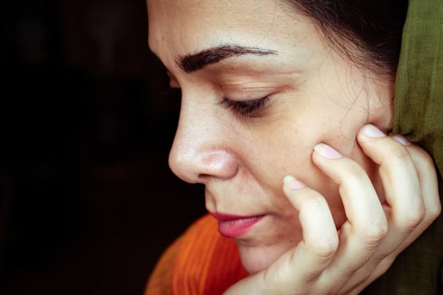 Close up profile portrait of a woman with dark hair and olive skin in a contemplative pose. She rests her chin gently on her hand with eyes downcast in reflection. She wears a green headscarf and orange accent clothing. The dark background emphasizes the peaceful, introspective quality of the image. Soft lighting highlights her thoughtful expression.