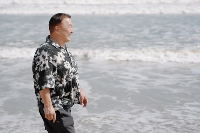 Middle aged man wearing a black and white floral patterned shirt standing at the edge of the ocean, looking peacefully toward the horizon. Gentle waves wash onto the sandy shore around his feet under an overcast sky