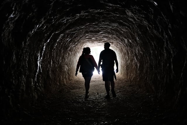 Silhouettes of two people walking side by side through a dark stone tunnel toward bright daylight at the exit. The tunnel walls have a rough, cave like texture. The figures appear to be a man and woman walking at a comfortable pace with natural light illuminating their destination ahead.