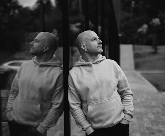 Black and white photograph of a bald man wearing a light colored hoodie, standing beside a large glass window or wall. He looks upward with a contemplative expression while his reflection is clearly visible in the glass. He has small earrings and his hands are in the hoodie pocket. The urban outdoor setting is softly blurred in the background. The image conveys introspection and self awareness.