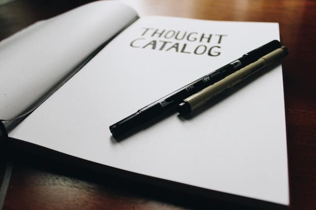 Open notebook or journal with "Thought Catalog" printed on the page, with two pens resting on top, placed on a dark wooden surface.