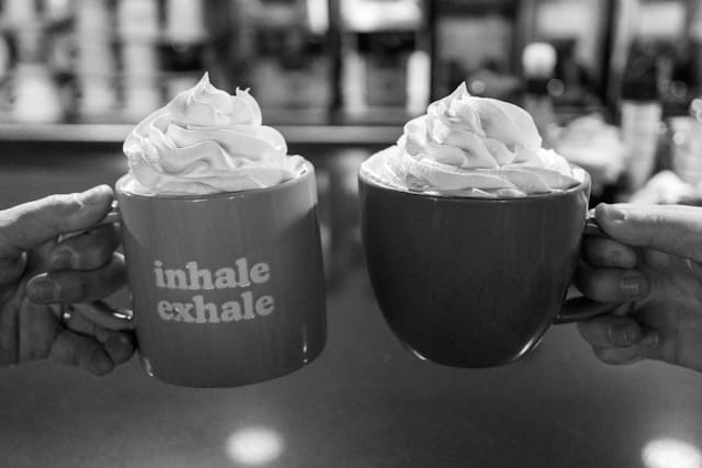 Two hands holding mugs topped with whipped cream, one mug reading inhale exhale, representing the power of breath in calming the nervous system