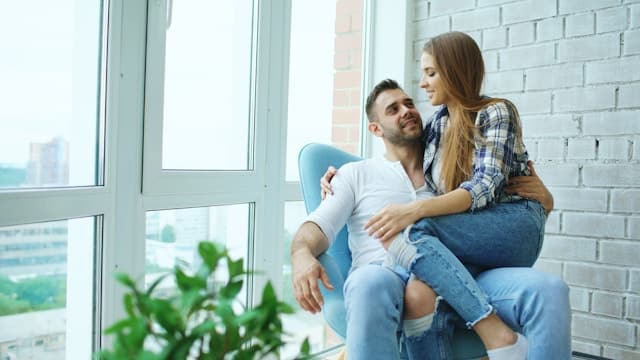 Couple sitting together by a bright window, looking at each other warmly with relaxed body language