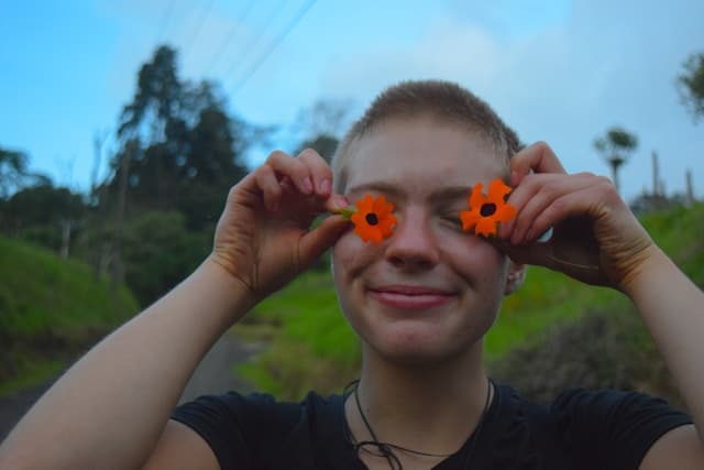 Person with short hair smiling playfully while holding two orange flowers over their eyes outdoors