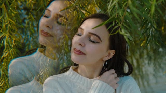 Woman with eyes closed beneath willow branches with a soft reflection beside her, representing inner awareness and the integration of mind and body in therapy