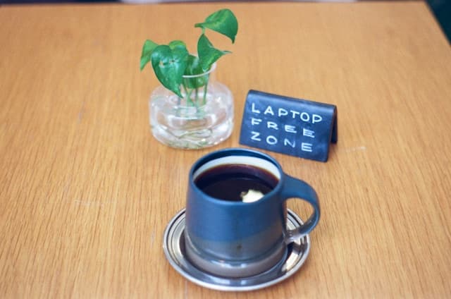 Cup of black coffee on a saucer beside a small plant and a sign reading Laptop Free Zone on a wooden table, representing the intentional pause and digital disconnection encouraged in mindfulness practice