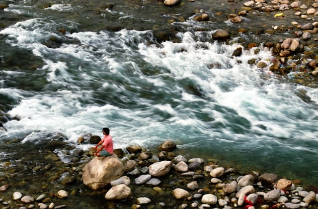 Person sitting quietly on a rock beside a rushing river, gazing at the flowing water, reflecting the stillness and present moment awareness cultivated in mindfulness therapy