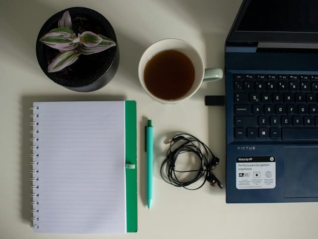 A flat lay view of a desk with a spiral notebook, green pen, cup of tea, small potted plant, wired earbuds, and a laptop computer