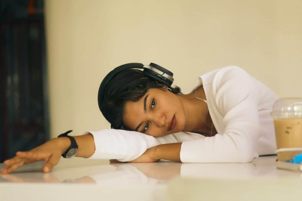 A young woman wearing over the ear headphones rests her head on her folded arms on a table, eyes open in a calm and reflective expression