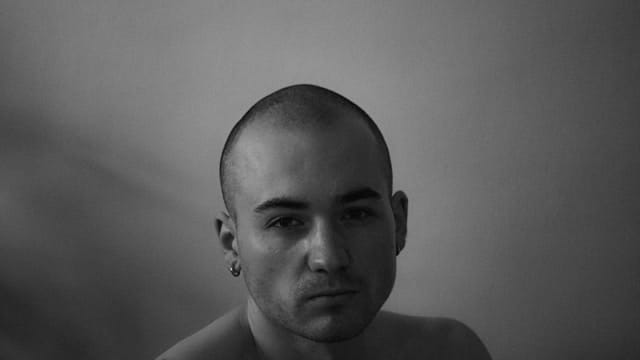Black and white portrait of person with shaved head and small earring looking directly at camera with thoughtful expression
