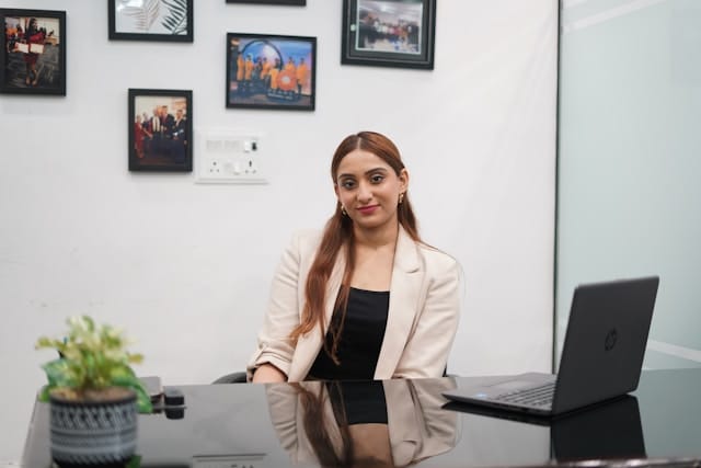 South Asian woman with long hair wearing cream blazer seated at desk with laptop, looking confidently at camera