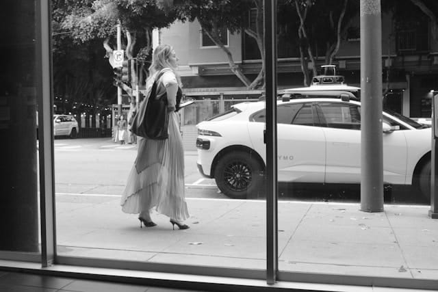 Black and white photograph of a woman in a flowing skirt walking along a San Francisco sidewalk, viewed through a glass storefront window with a Waymo autonomous vehicle visible on the street, evoking the texture of Bay Area urban life.